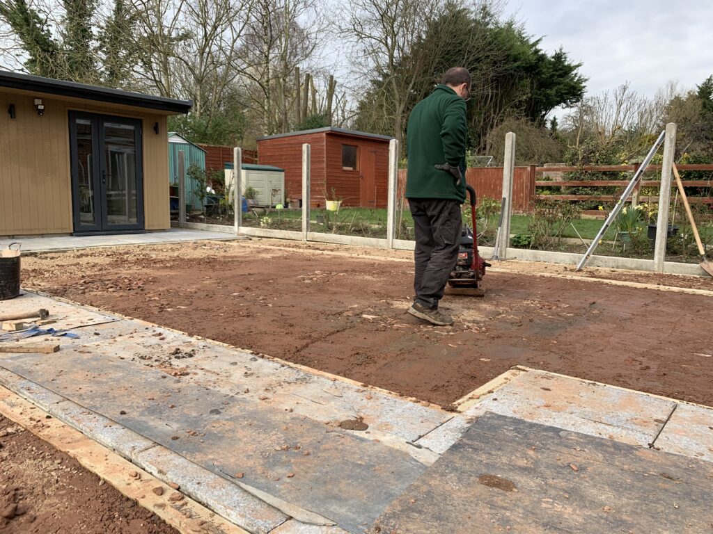 Gardener preparing the ground for artificial grass installation, levelling soil and compacting the base in a residential garden