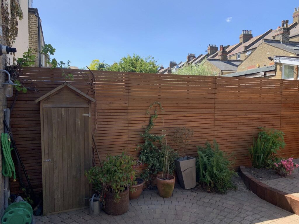 Modern London garden with horizontal wooden fence panels, garden shed, paved patio area, and planted borders, showing stylish fencing and small garden design